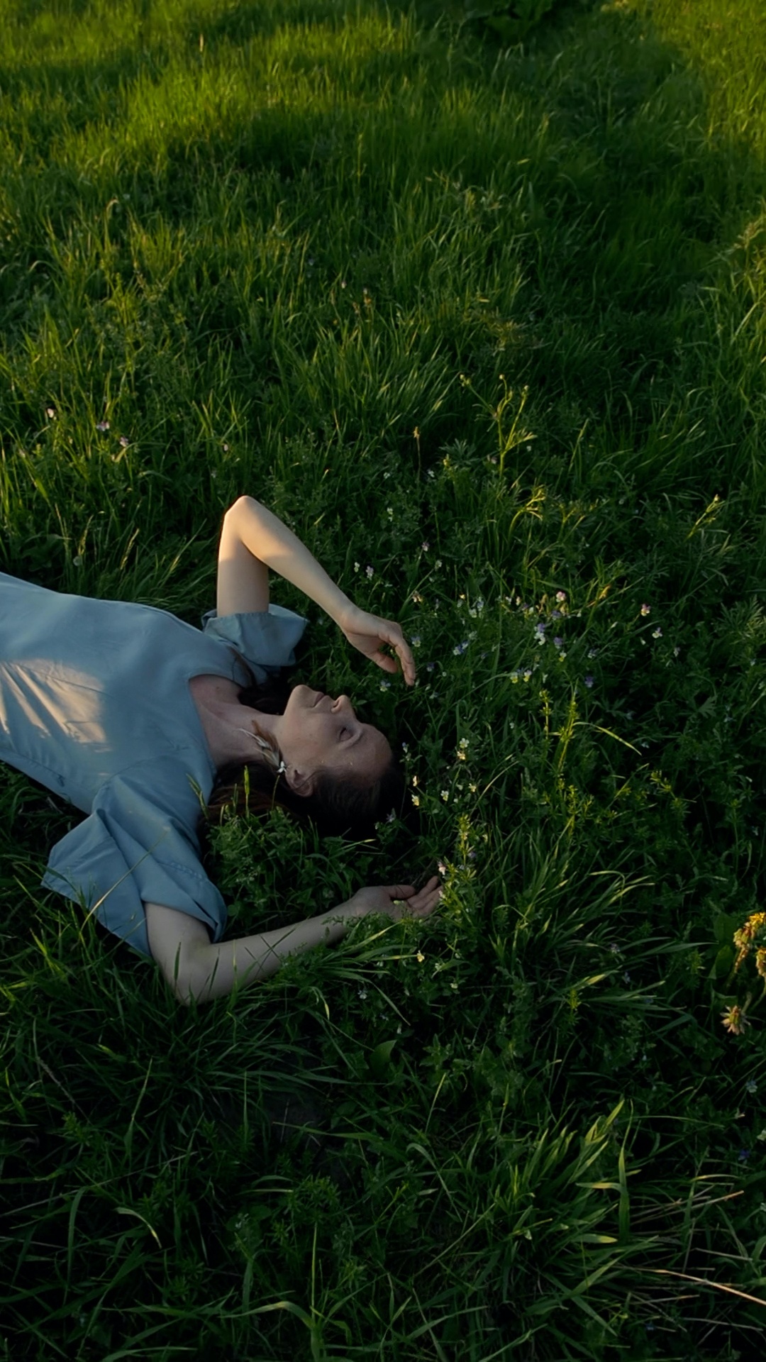 A woman lies motionless in a lush green meadow during golden hour. She remains completely still, ...