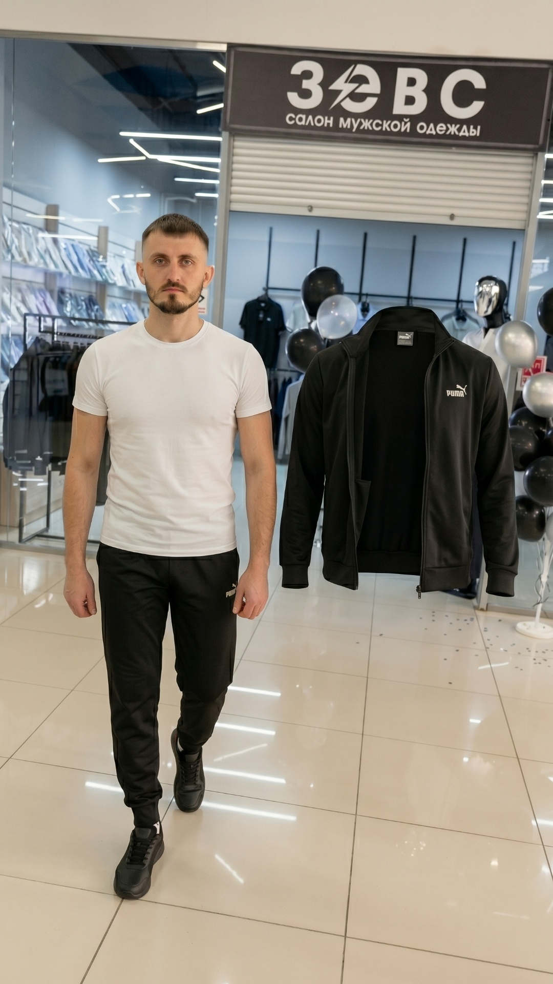 A man wearing a white t-shirt and black track pants is walking forward inside a clothing store. T...