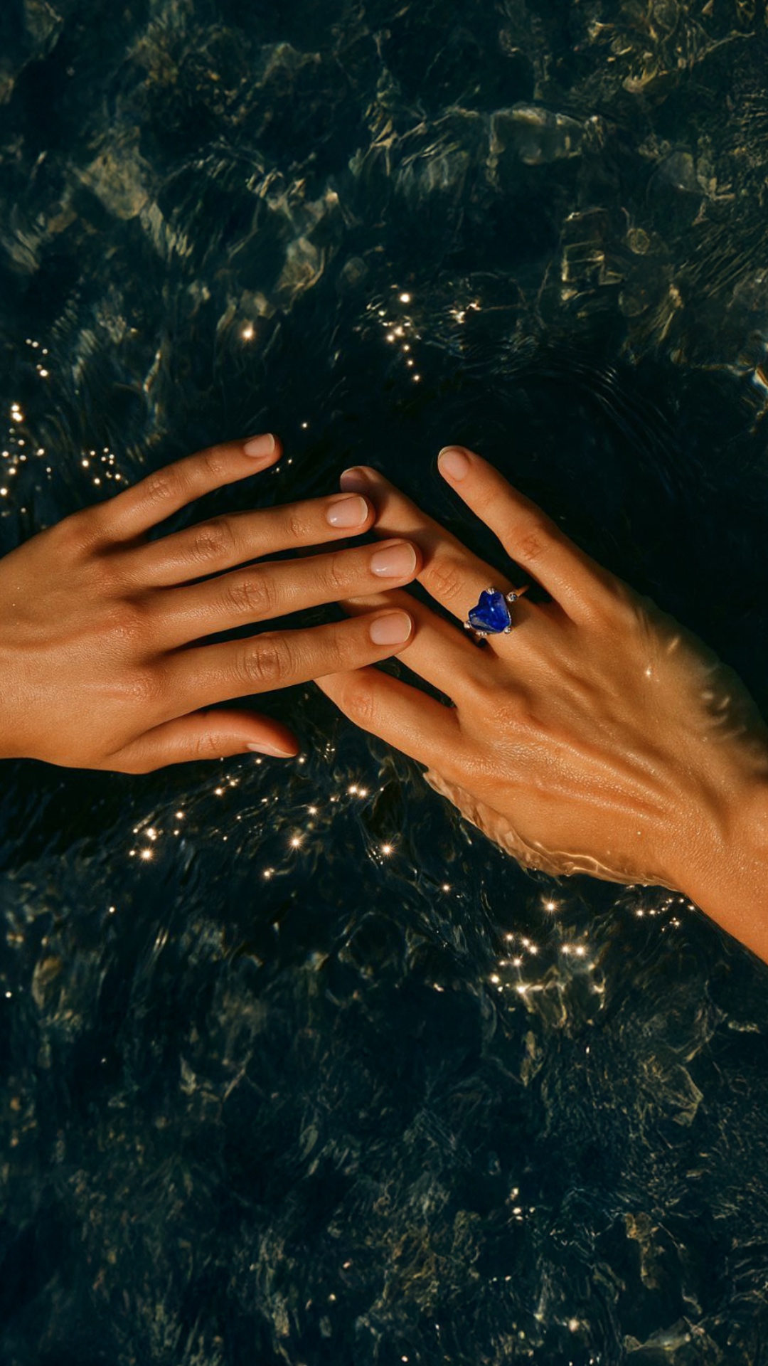 Subject: Two elegant female hands, one adorned with a silver ring featuring a vivid blue heart-sh...