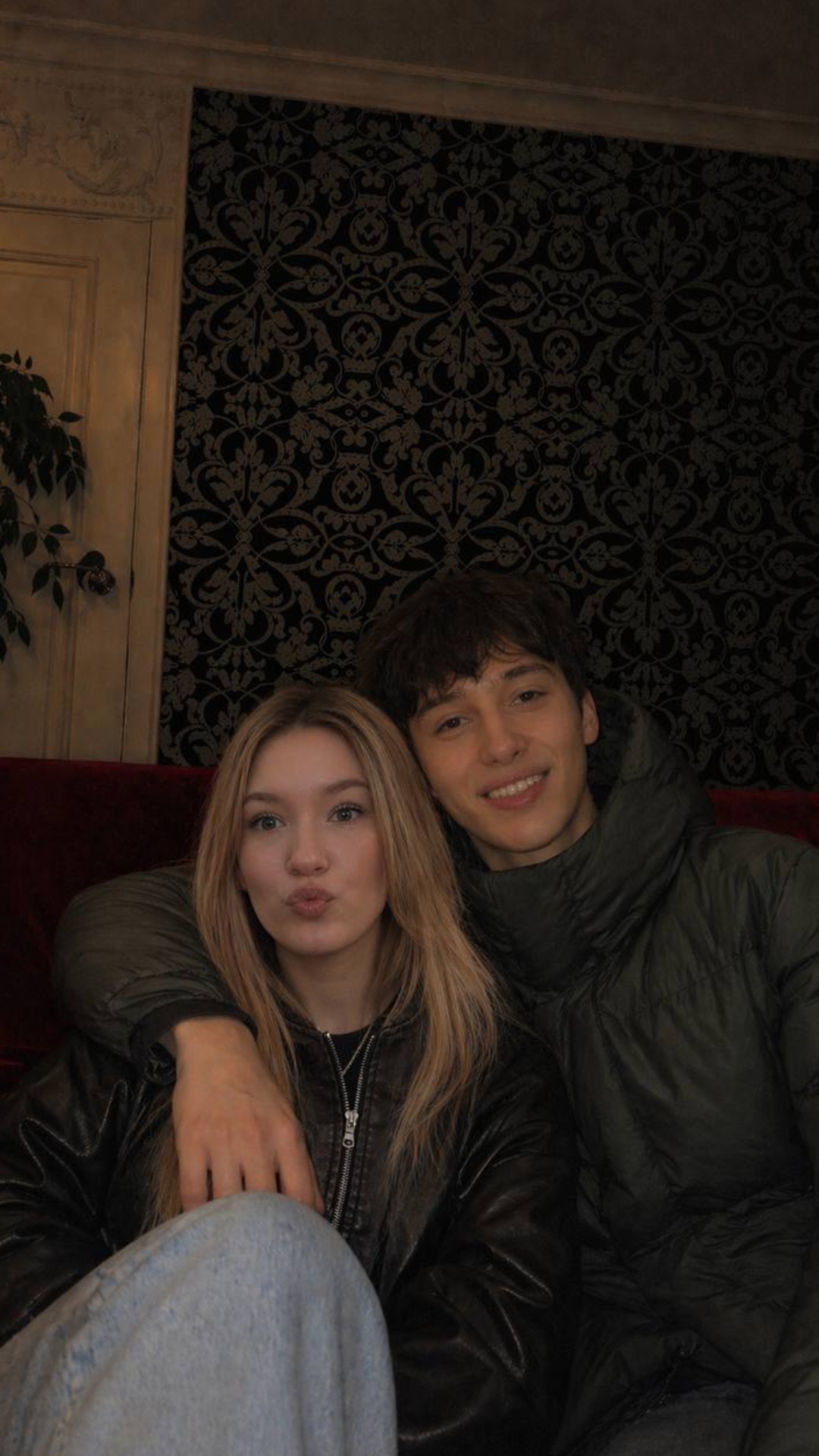 A young couple sitting very close together on a red velvet couch in a dimly lit cozy room with da...